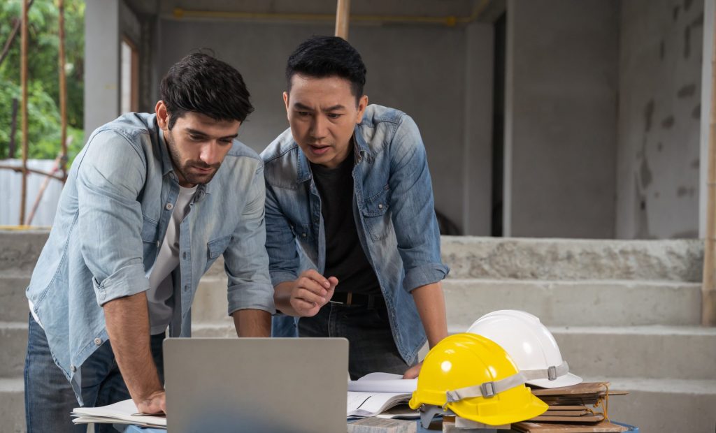 Two people on a building site are talking and looking at a laptop. Two hard hats are on the table next to them.