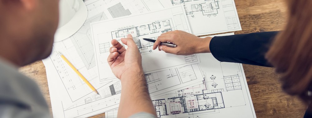 Two people point to different rooms in a blueprint of a floor plan on the wooden table in front of them.