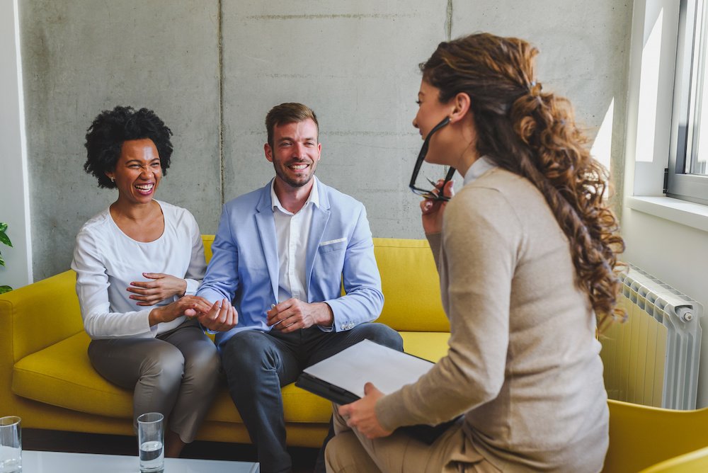 A smiling couple sit on a yellow sofa talking to their interior designer in their first meeting.