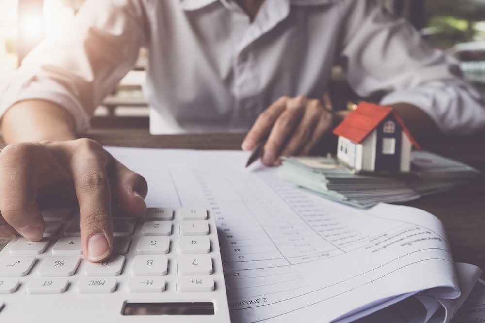 A person sits at a desk with a small model home and money next to them to illustrate the costs of buying a home. They use a calculator to add up costs.