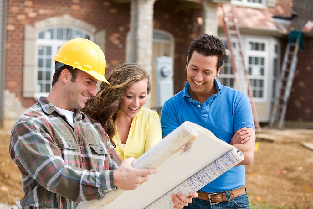A happy couple stands with a builder looking at the blueprint for their future home as it's being built behind them.