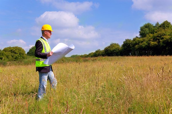 An architect wearing a hard hat and high-vis jacket assesses a greenfield site to build a property there.