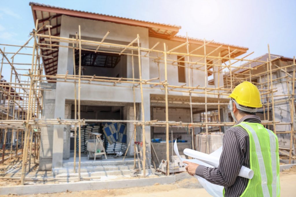 An engineer in a hard hat and hi-vis stands with blueprints looking at a home under construction, surrounded by scaffolding.