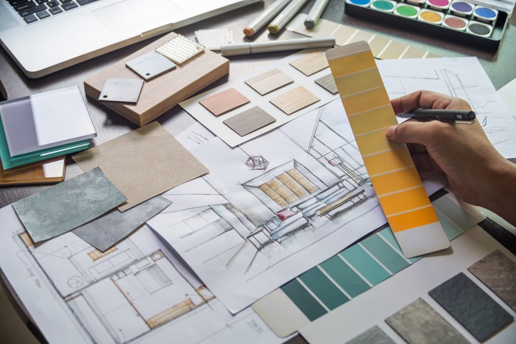 Interior designer sits at their desk with a spread of paint, tile, and fabric swatches in front of them. There's also a drawing of a living room space and a blueprint of a floor plan.