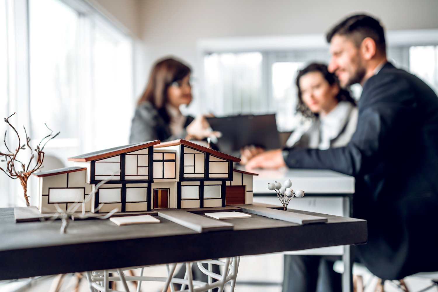 A model home sits on a desk in the foreground, while the homeowners-to-be talk to an advisor in the background.